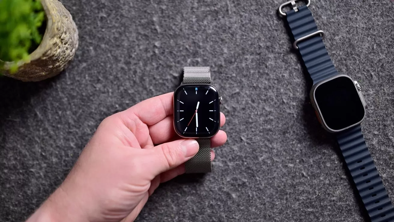 Hand holding a modern square smartwatch with a metal band, displaying an analog-style watch face, next to another similar smartwatch with a dark striped band on a gray fabric surface