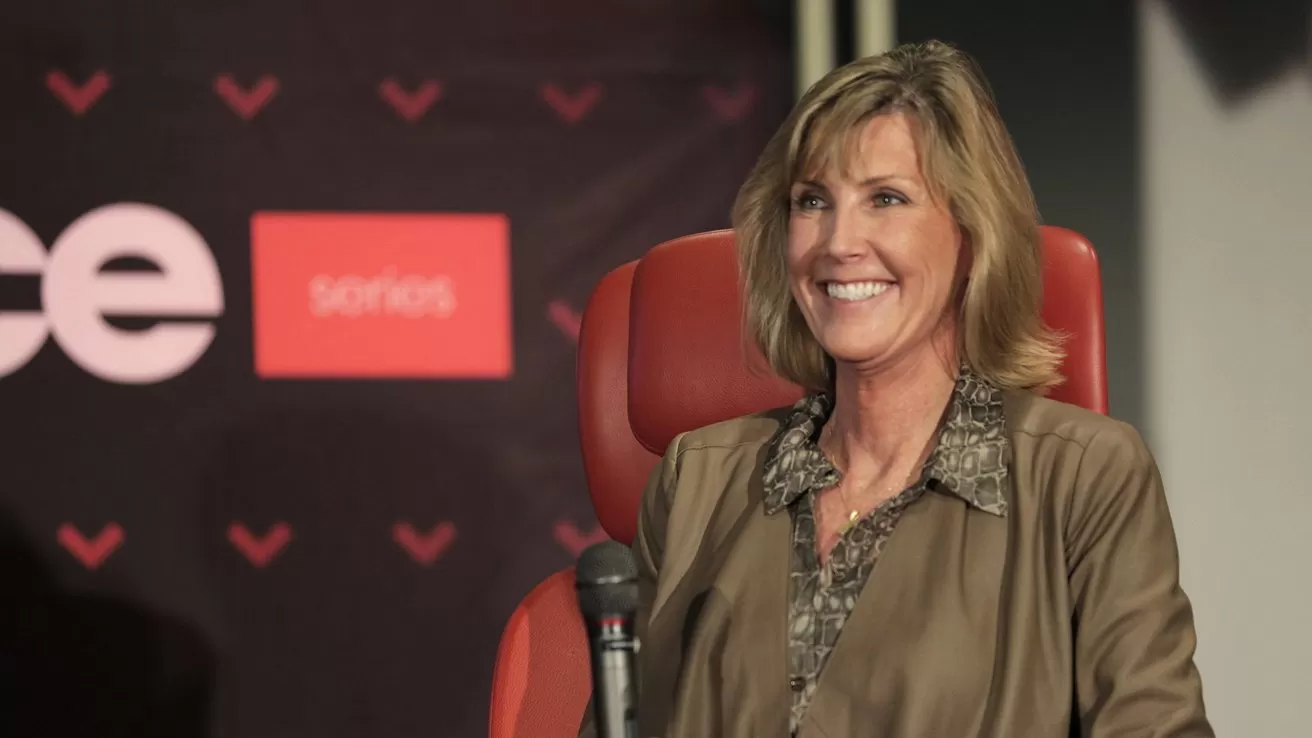 Smiling woman with short blond hair sits in a red chair on a stage, wearing a brown jacket, with a microphone in front and a conference backdrop behind her