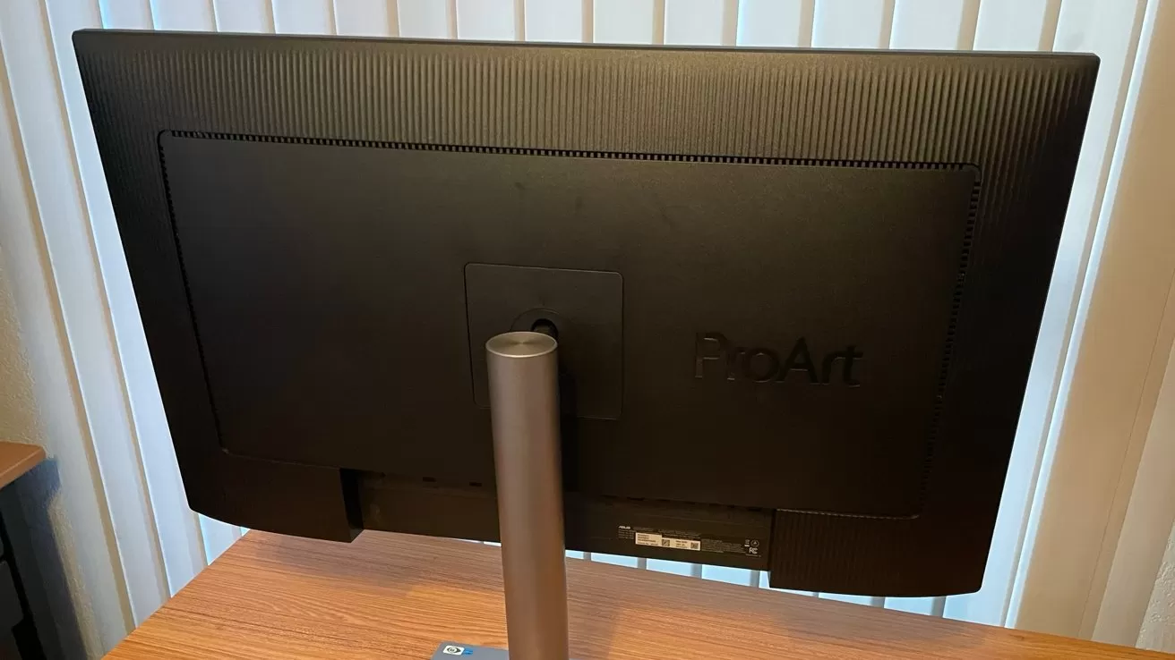 Back view of a black computer monitor labeled ProArt on a wooden desk, with vertical blinds in the background.
