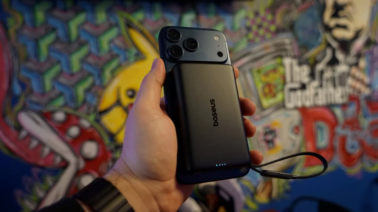 Hand holding an iPhone 17 Pro Max attached to a black Baseus power bank with glowing indicator lights, against a colorful graffiti-style mural featuring cartoon characters and bold text