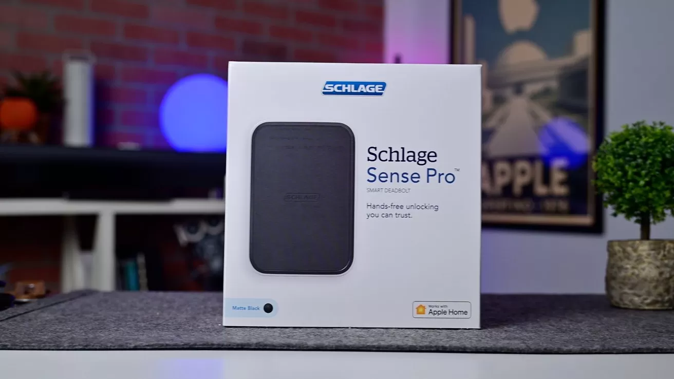 Box of Schlage Sense Pro smart lock on a table in a modern living room, with blurred shelves, decorations, and framed wall art in the background