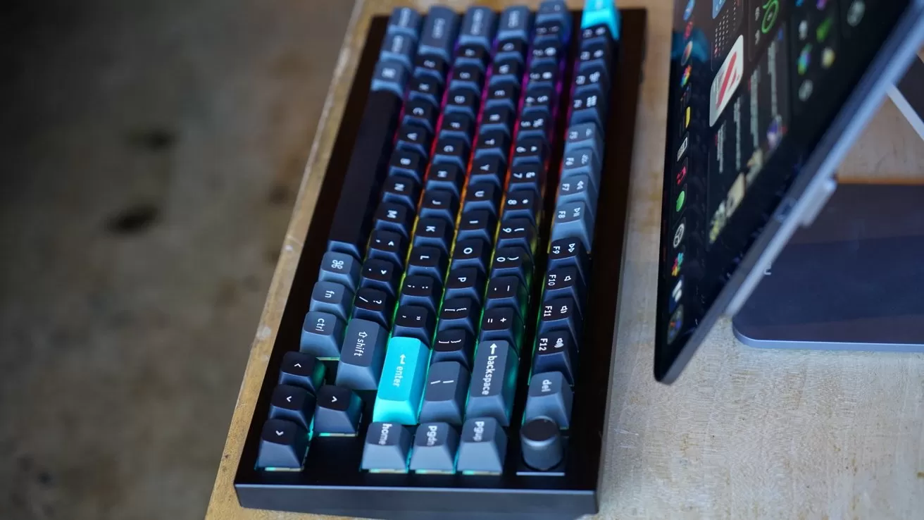 Mechanical keyboard with dark keycaps and colorful RGB backlighting on a wooden desk, next to a tablet or laptop on a stand displaying app icons and text