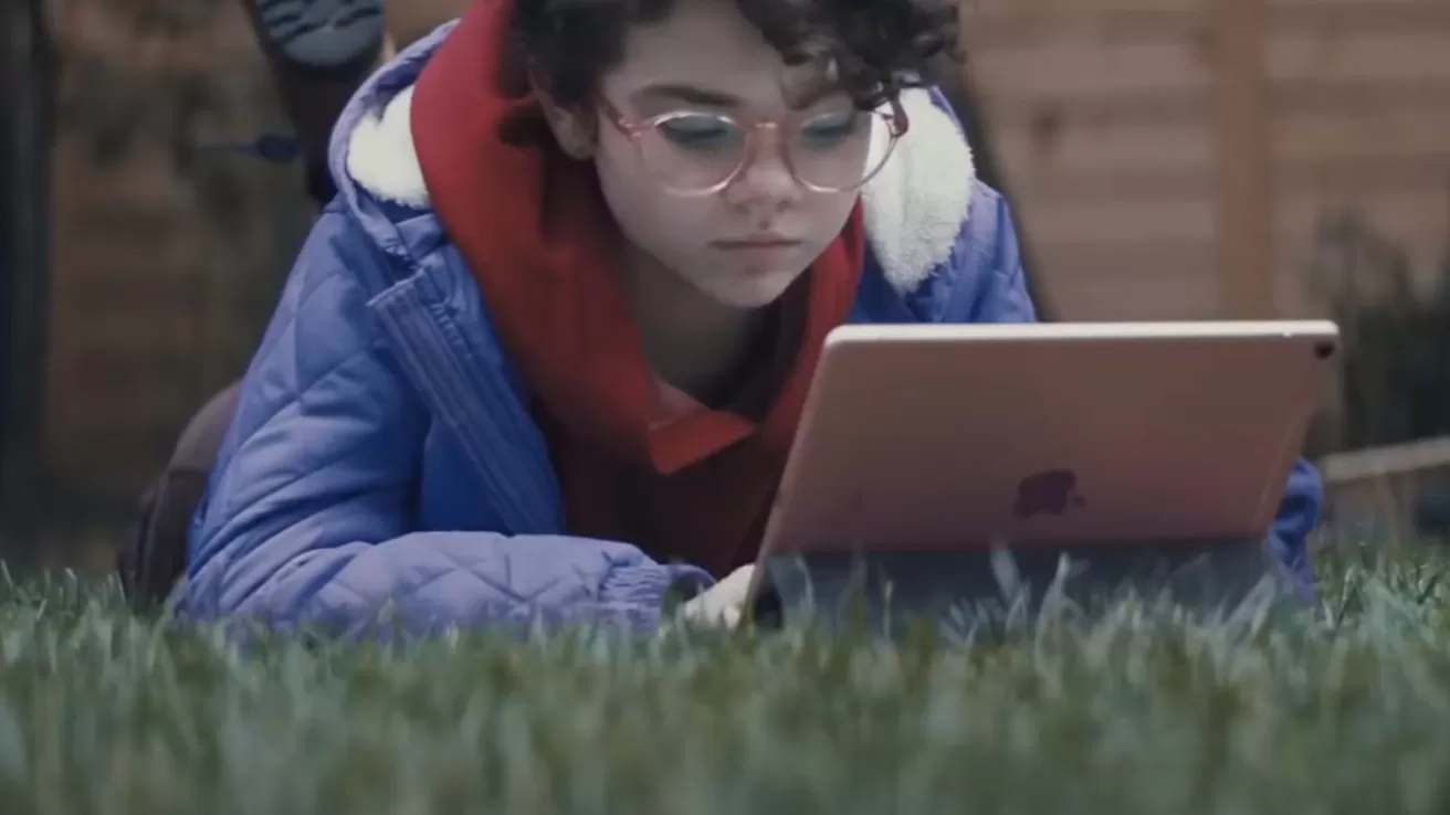 Child with curly hair and glasses lying on grass outdoors, wearing a blue jacket and red hoodie, intently using a tablet computer in a backyard setting