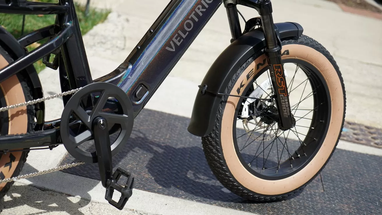 Closeup of a Velotric electric bicycle showing crank, chain, and large front wheel with tan Kenda tire, black fender, and suspension fork on a sunny sidewalk.