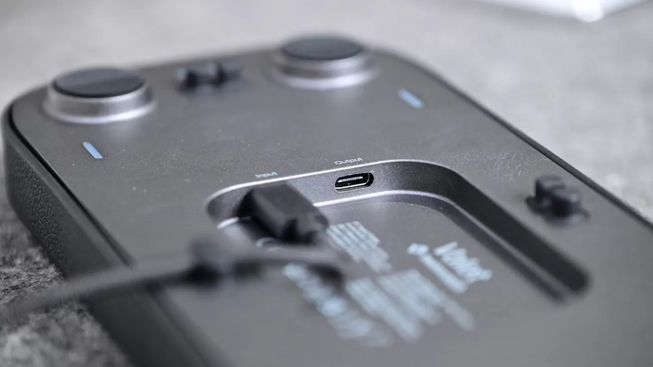 Close-up of a dark handheld gaming controller lying flat, showing a USB-C charging port with a cable partially plugged in, two joystick-like controls, and small buttons on a gray surface