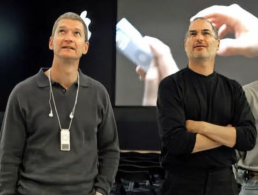 Two men stand side by side at a tech event, looking upward thoughtfully, with a large screen behind them showing hands holding a small electronic device.