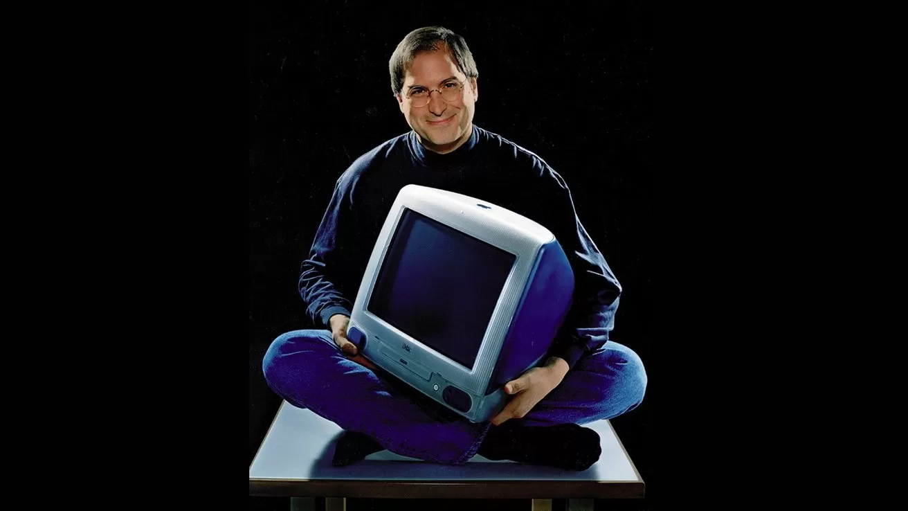 Smiling man in glasses and dark turtleneck sits cross-legged on a table, holding a colorful translucent CRT desktop computer against a dark background