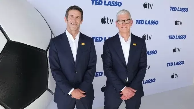 Two men in dark suits stand smiling on a red carpet beside a giant soccer ball, with a backdrop featuring the Ted Lasso logo and Apple TV branding