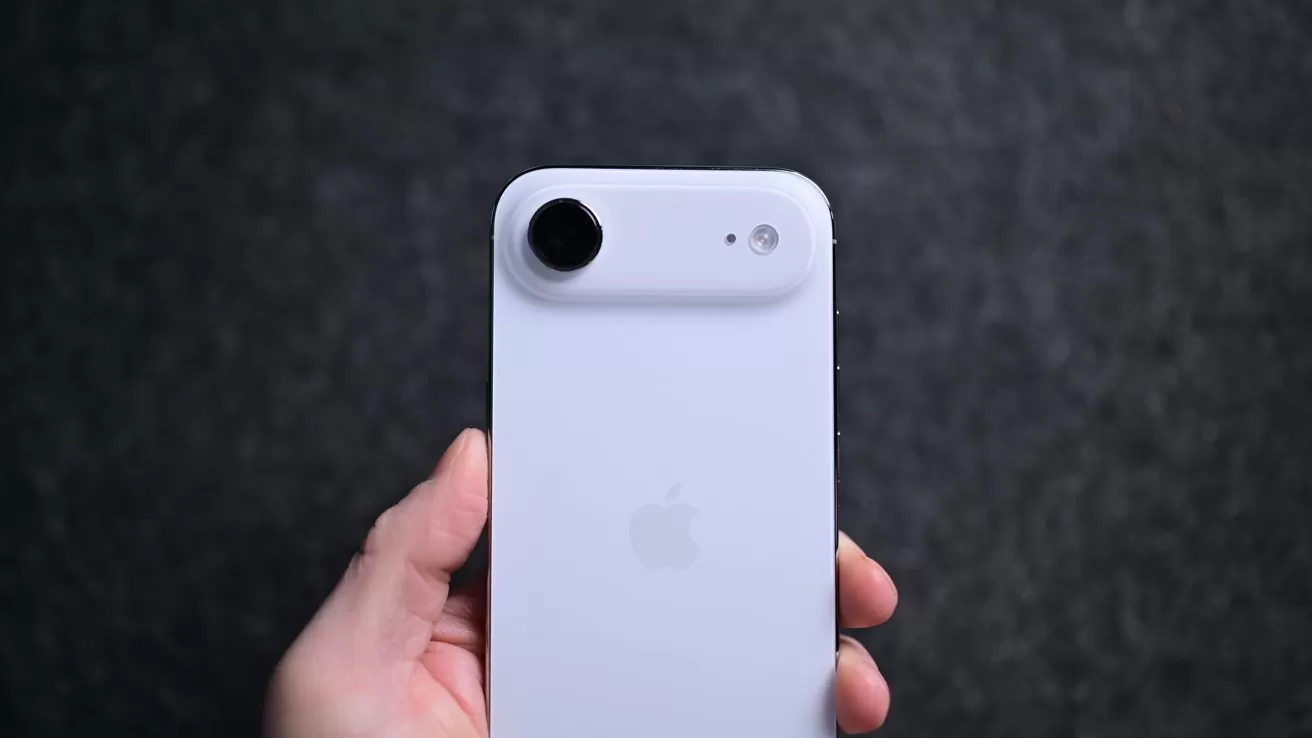 Hand holding a white iPhone seen from the back, featuring a large circular camera lens attachment on the top left against a dark, blurred background