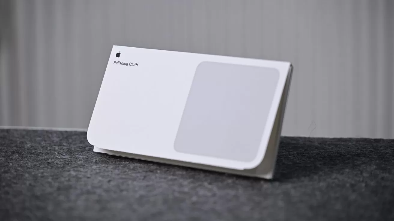 White Apple polishing cloth in folded packaging, standing upright on a dark felt surface with a gray vertically striped background