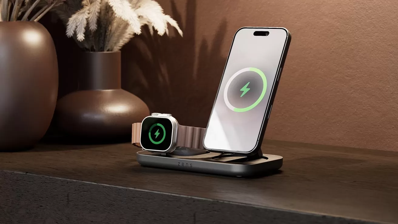 Smartphone and smartwatch charging on a sleek wireless dock, both screens showing green charging symbols, placed on a dark tabletop with blurred decorative vases and dried plants in the background