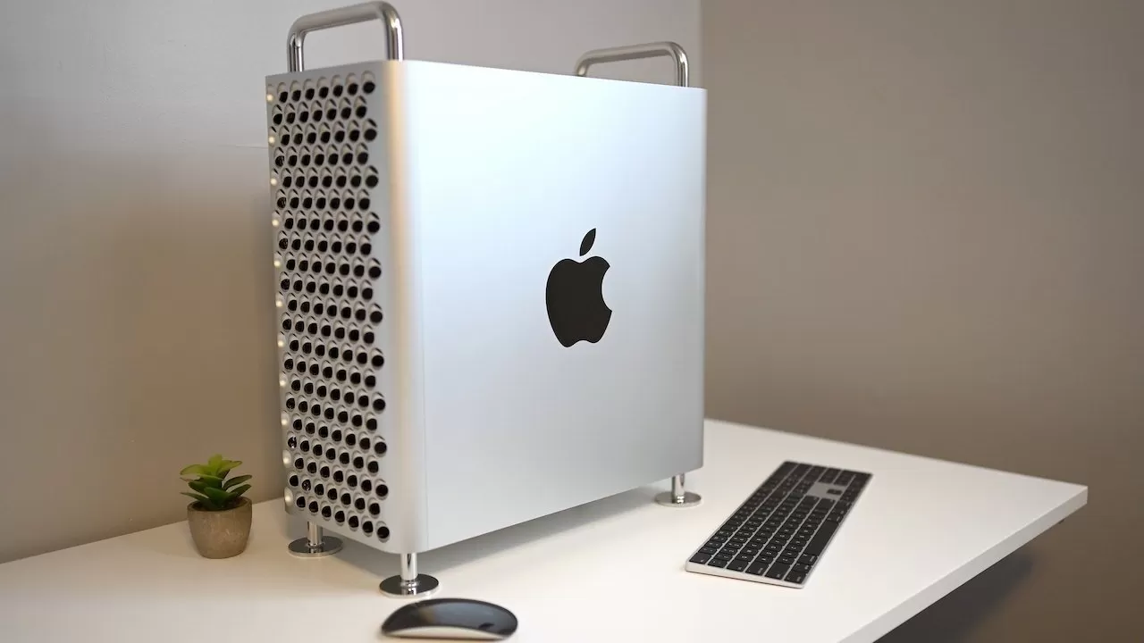 Silver desktop computer with a perforated front and Apple logo, on a white desk, accompanied by a keyboard, mouse, and small potted plant.