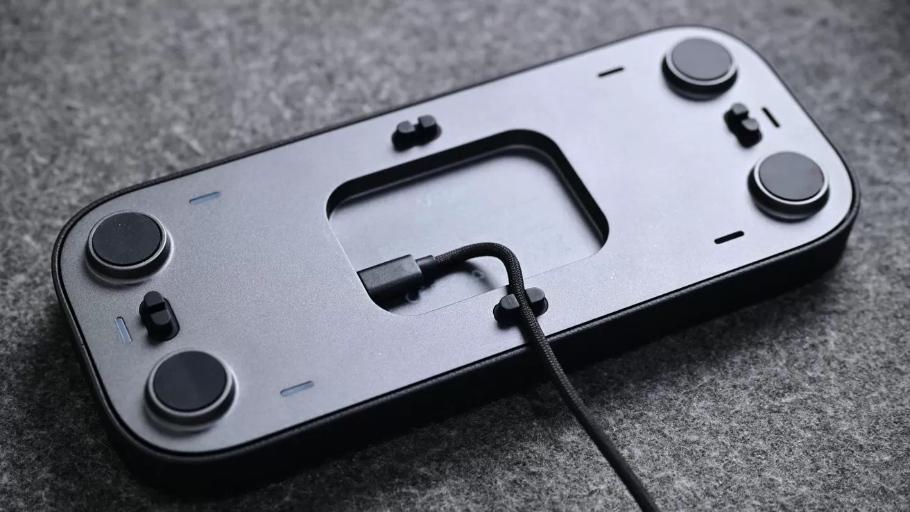 Flat rectangular electronic device with four round rubber feet on the underside, resting on gray fabric, featuring a central cutout and a single cable plugged into a port in the middle