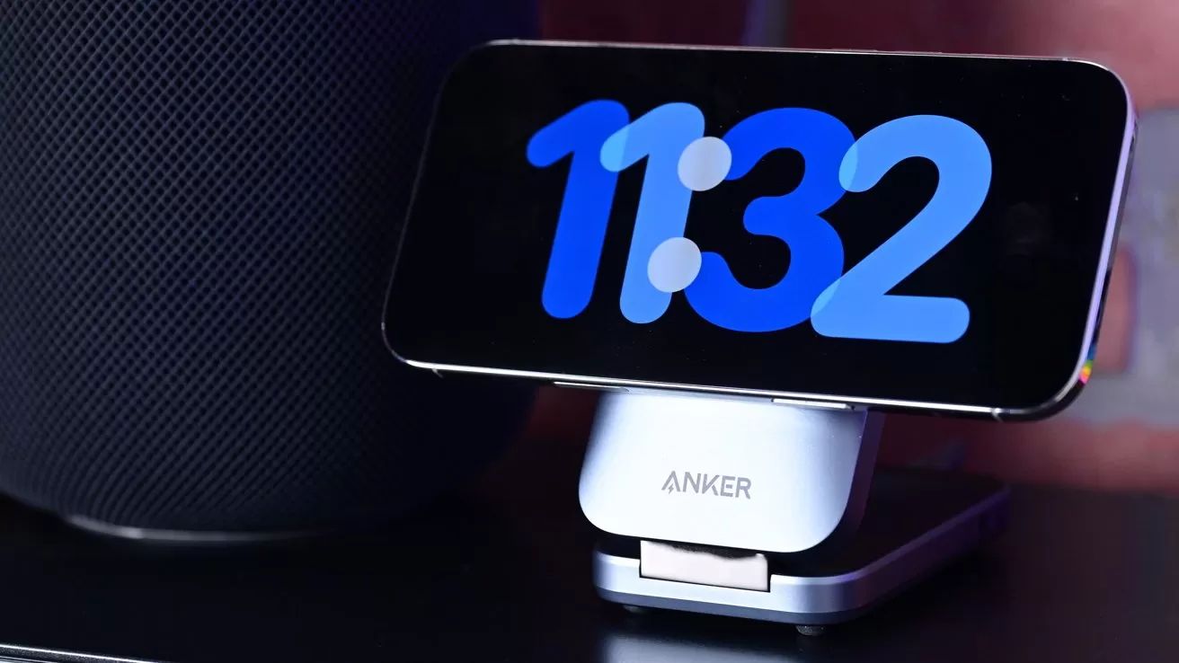 Smartphone on an Anker charging stand displaying a large blue digital clock reading 11:32, placed on a dark surface beside a tall cylindrical speaker