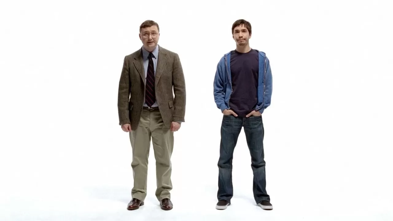 Two men stand against a plain white background, one dressed formally in a brown blazer and tie, the other casually in a blue hoodie and jeans, both facing forward