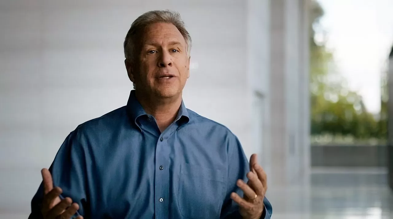 Middle-aged man in a blue button-down shirt speaking with hands raised slightly, standing indoors near large windows with soft natural light and blurred greenery in the background