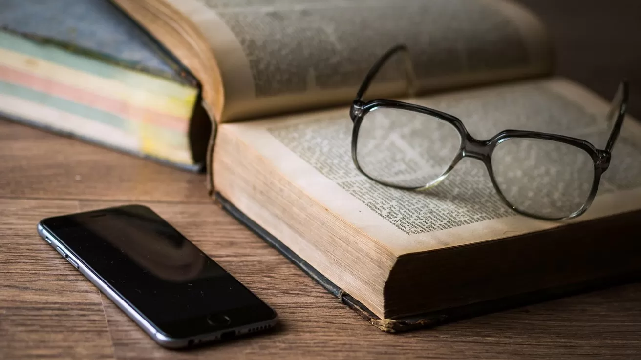 Open book on wooden table with reading glasses resting on its pages, a smartphone lying beside it, and another closed book stacked in the background