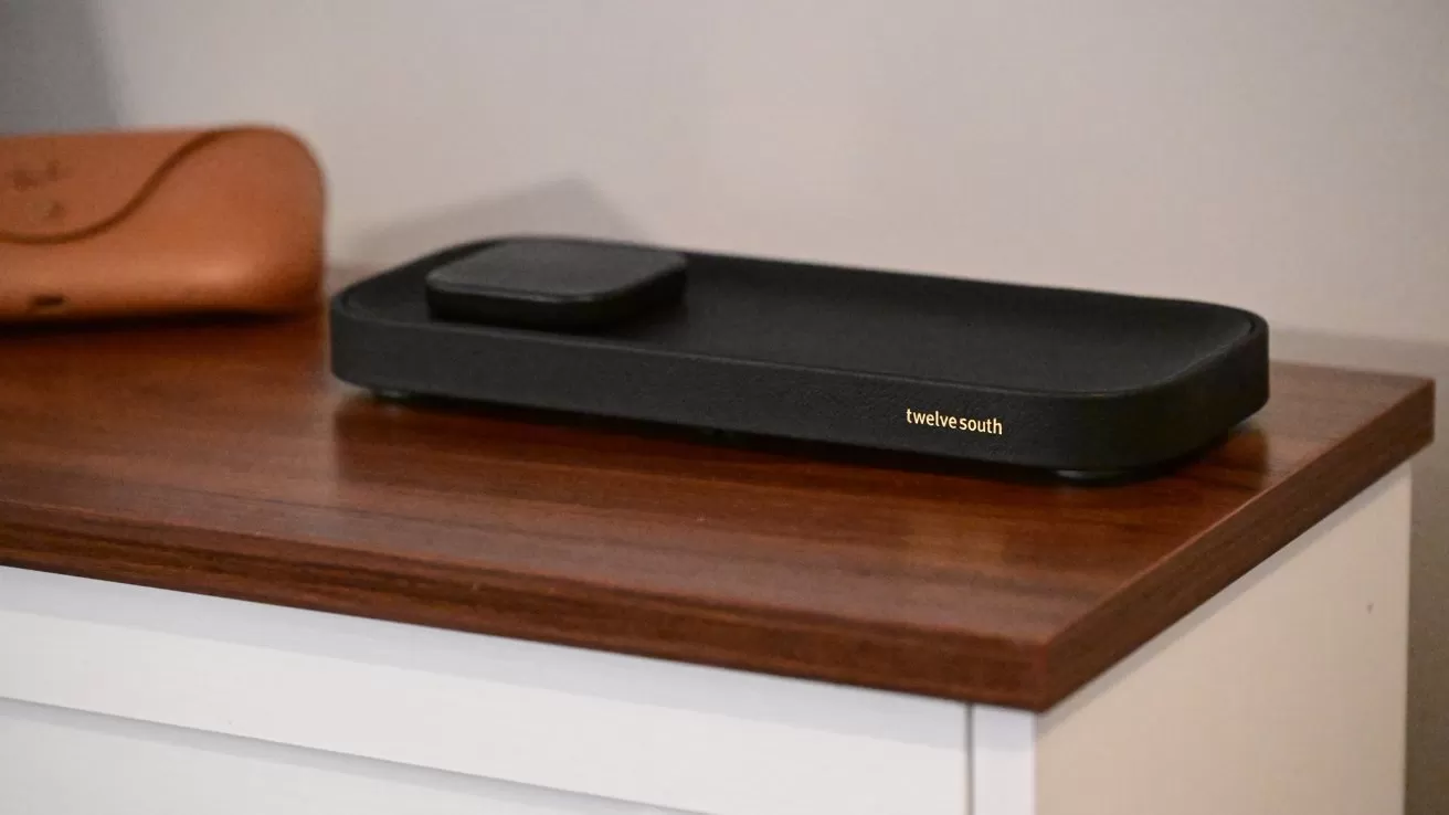 Black Twelve South charging dock with a small raised pad sits on a wooden-topped white cabinet, with a tan leather case placed nearby in a softly lit room