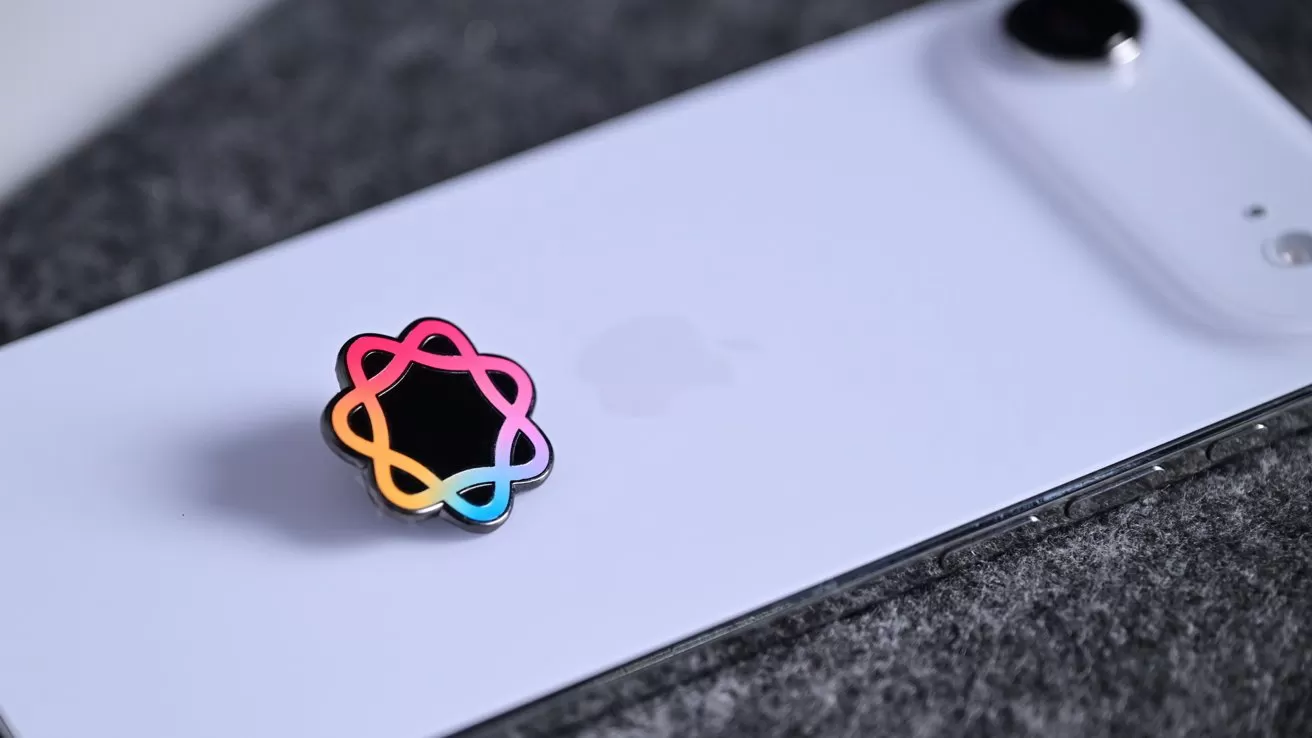 White smartphone lying face down on dark fabric, featuring a black circular pin with a colorful intertwined outline in pink, orange, yellow, green, and blue near the Apple logo