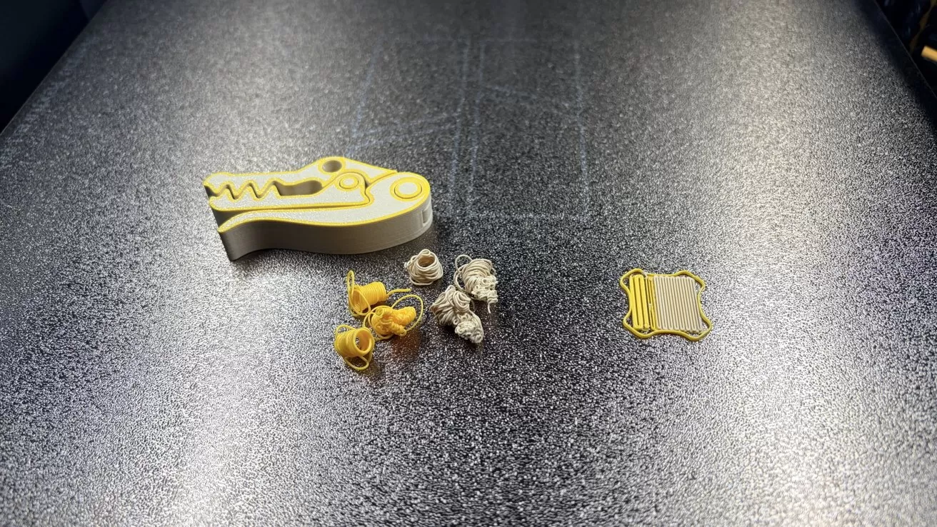 3D printer bed with a completed yellow and white crocodile-shaped part, several tangled filament blobs in yellow and white, and a small flat rectangular yellow frame on the right
