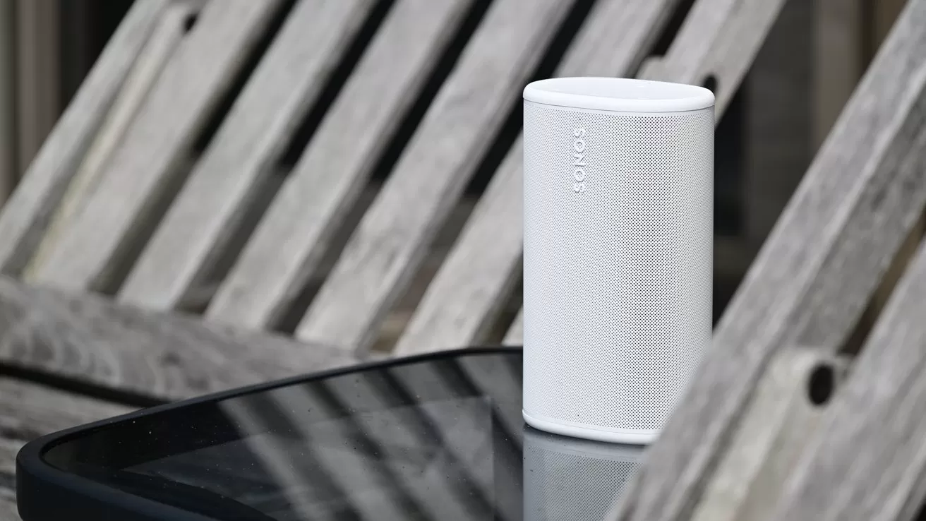 White Sonos wireless speaker standing on a reflective black table outdoors, with weathered wooden slats of a bench in the background
