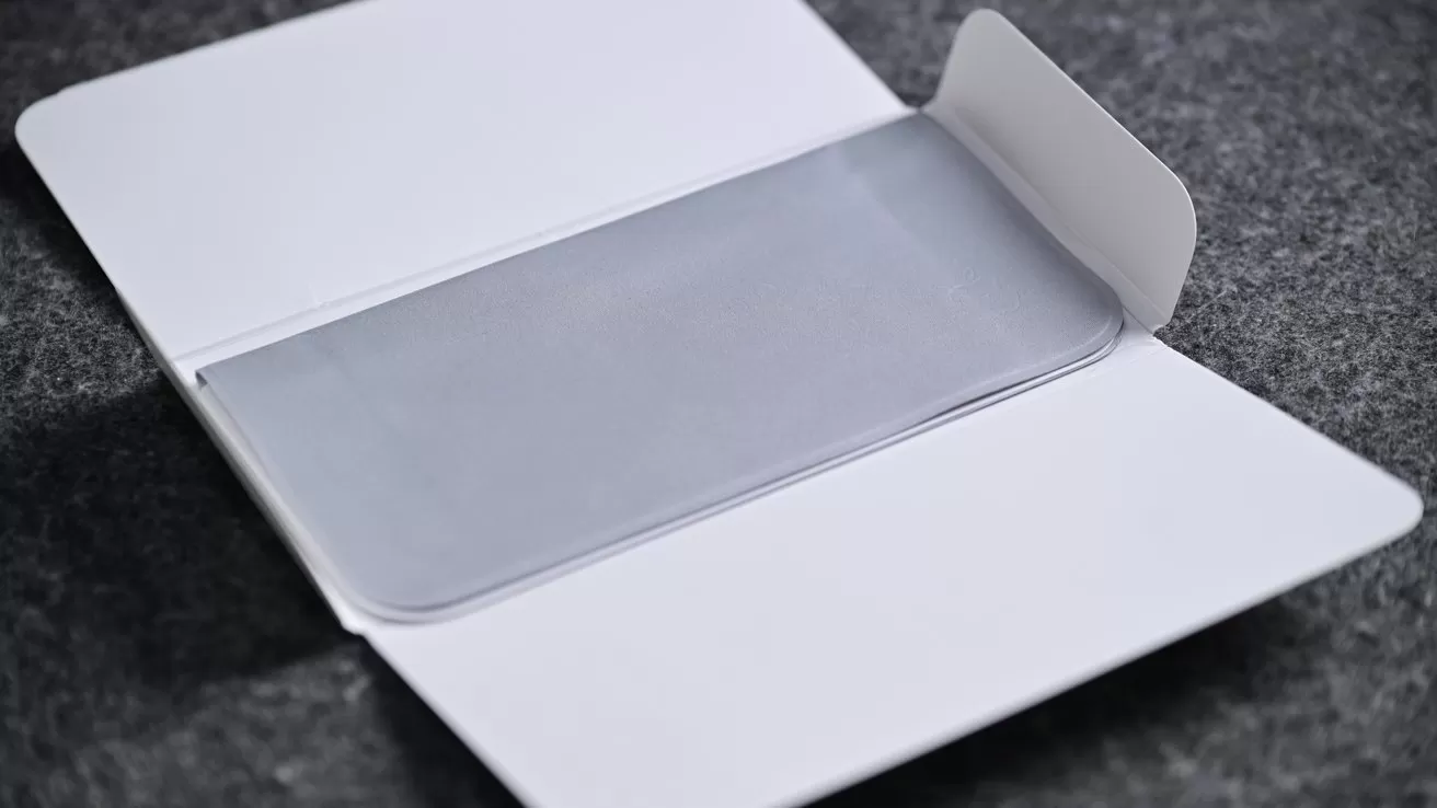 White cardboard packaging lying open on a dark surface, revealing a slim, translucent protective sleeve or film neatly centered inside the unfolded flaps