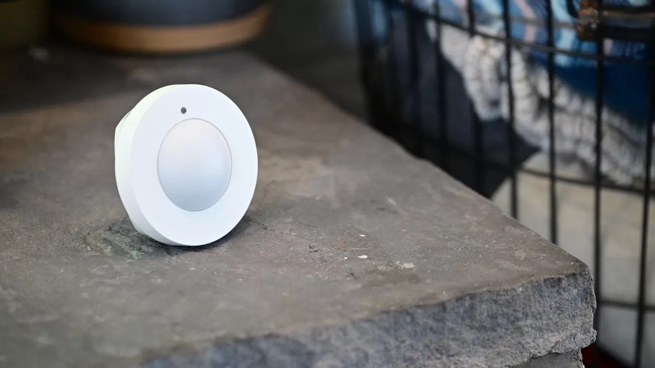 Small white circular smart sensor with a central dome resting on a rough stone surface, with a wire storage basket and blurred household objects in the background
