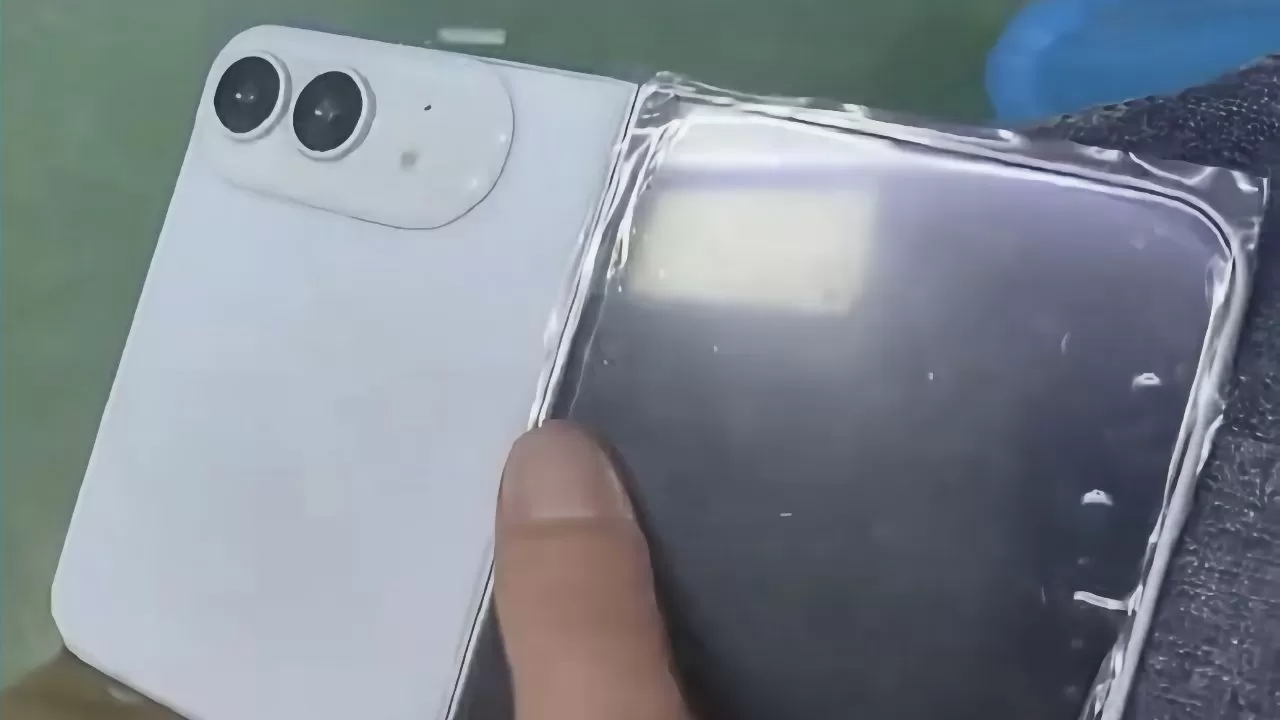 Hand holding a white foldable smartphone, partly open, showing dual rear cameras and a reflective outer screen, resting on someone's jeans with a blurred background.