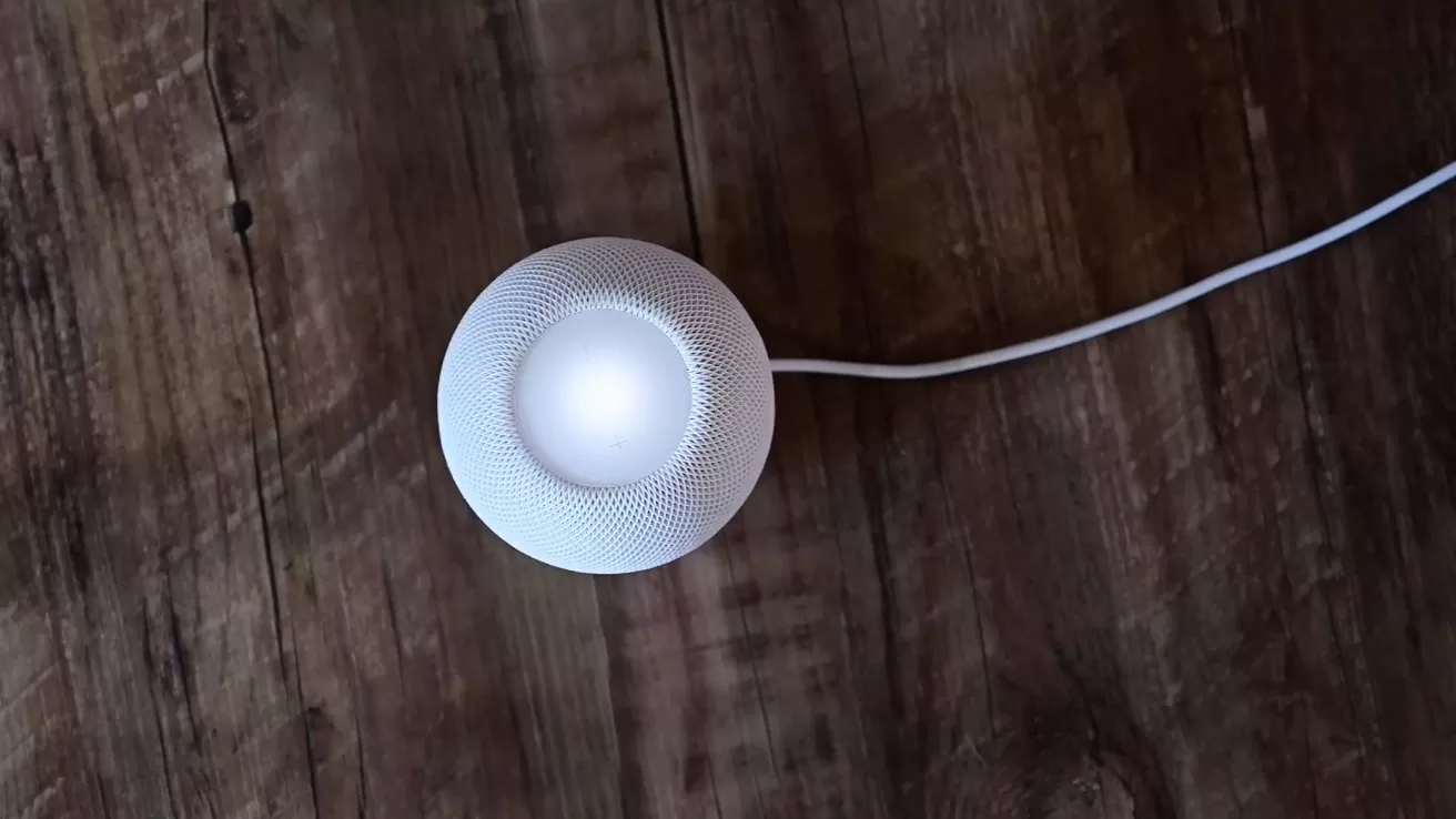 Small white smart speaker with glowing center sits on rustic wooden surface, connected by a thin white power cable extending to the right