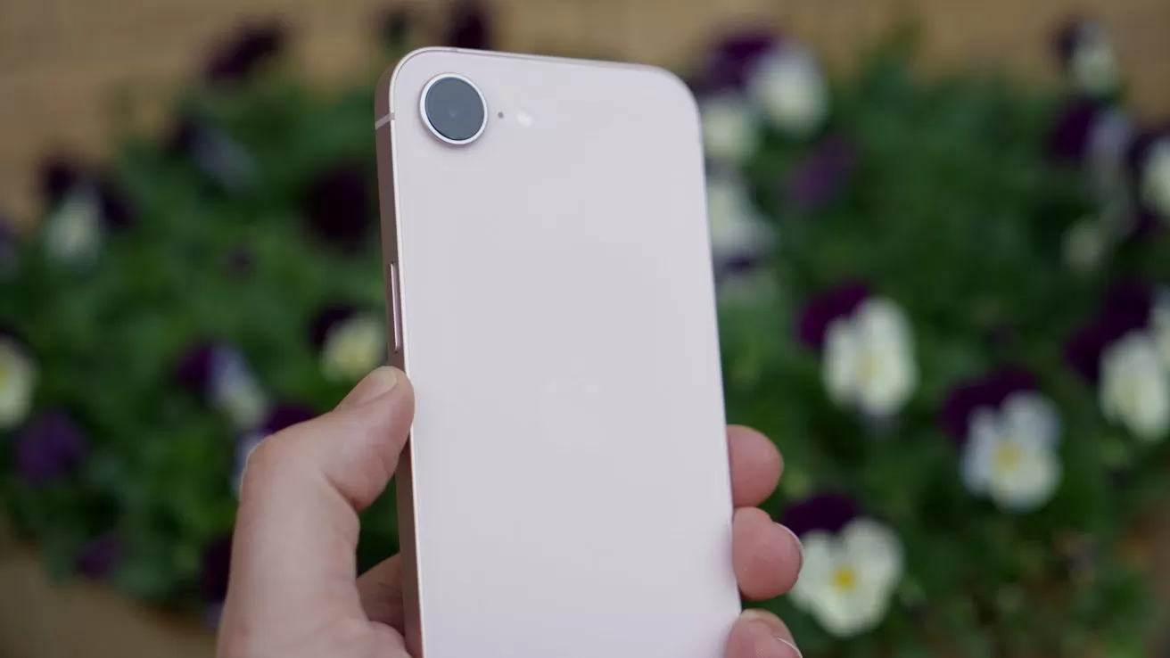 Hand holding a light pink smartphone showing its back, with a single rear camera, against a blurred background of green foliage and purple and white flowers