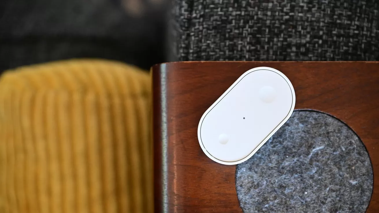 Small white oval smart sensor on a wooden furniture edge beside a gray felt pad, with textured gray upholstery above and mustard yellow corduroy fabric in the background