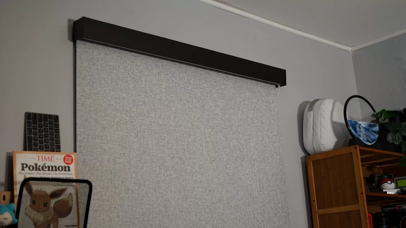 Room corner with a large gray pull-down blind, wooden shelves holding books and decor, a white backpack hanging on the side, and Pokemon magazine partially visible on the left
