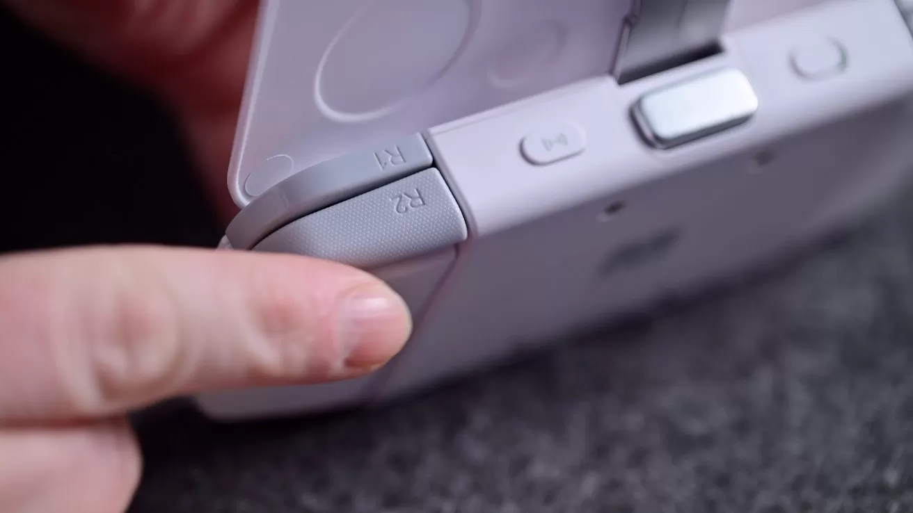 A person presses the R2 shoulder button on a handheld gaming console, showing a close-up of the controller's back.
