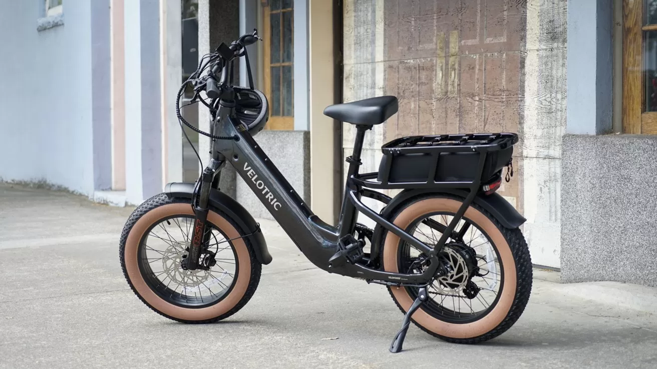 Black Velotric electric bike with tan fat tires, step-through frame, rear cargo rack, and kickstand, parked on a sidewalk in front of a building with windows and textured walls