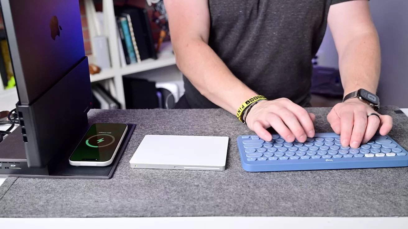 Person typing on a blue wireless keyboard at a tidy desk with a smartphone on a charging pad, a trackpad, a vertical laptop stand, and shelves of books in the background
