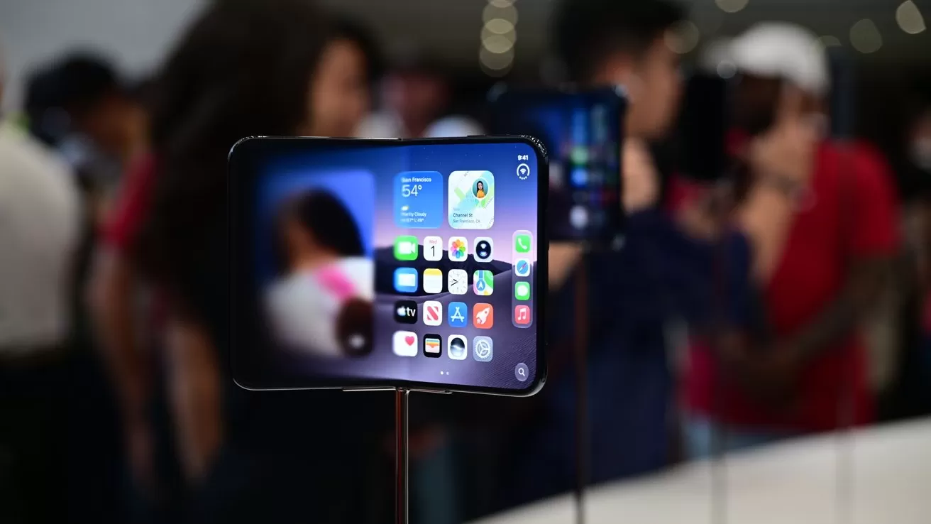 Smartphone on display stand showing colorful home screen icons and widgets, sharply focused in foreground, with a blurred crowd of people and bright indoor lighting in the background