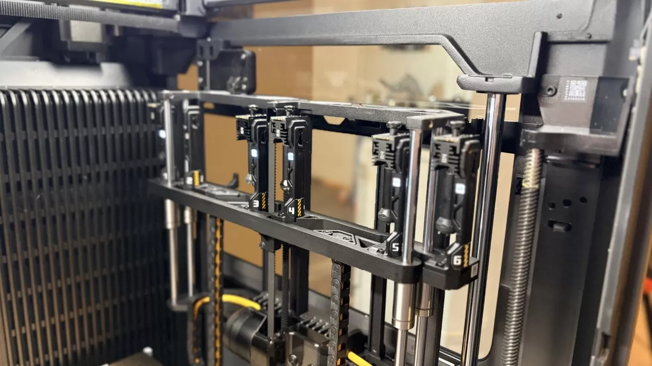 Close-up of an industrial 3D printer interior showing numbered toolheads on a metal gantry, vertical guide rods, belts, wiring, and mechanical components inside a partially enclosed frame