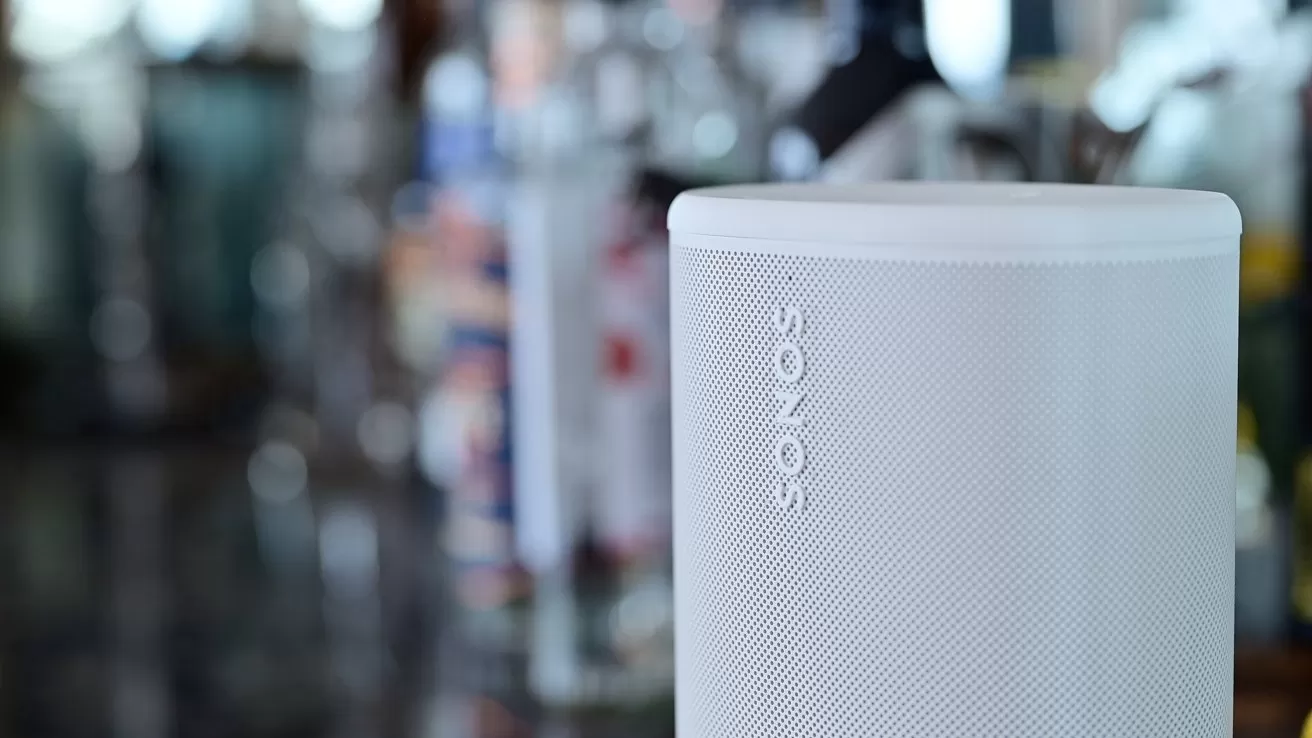 Close-up of a white Sonos wireless speaker on a reflective surface, with softly blurred bottles and bar-like background suggesting a modern indoor setting