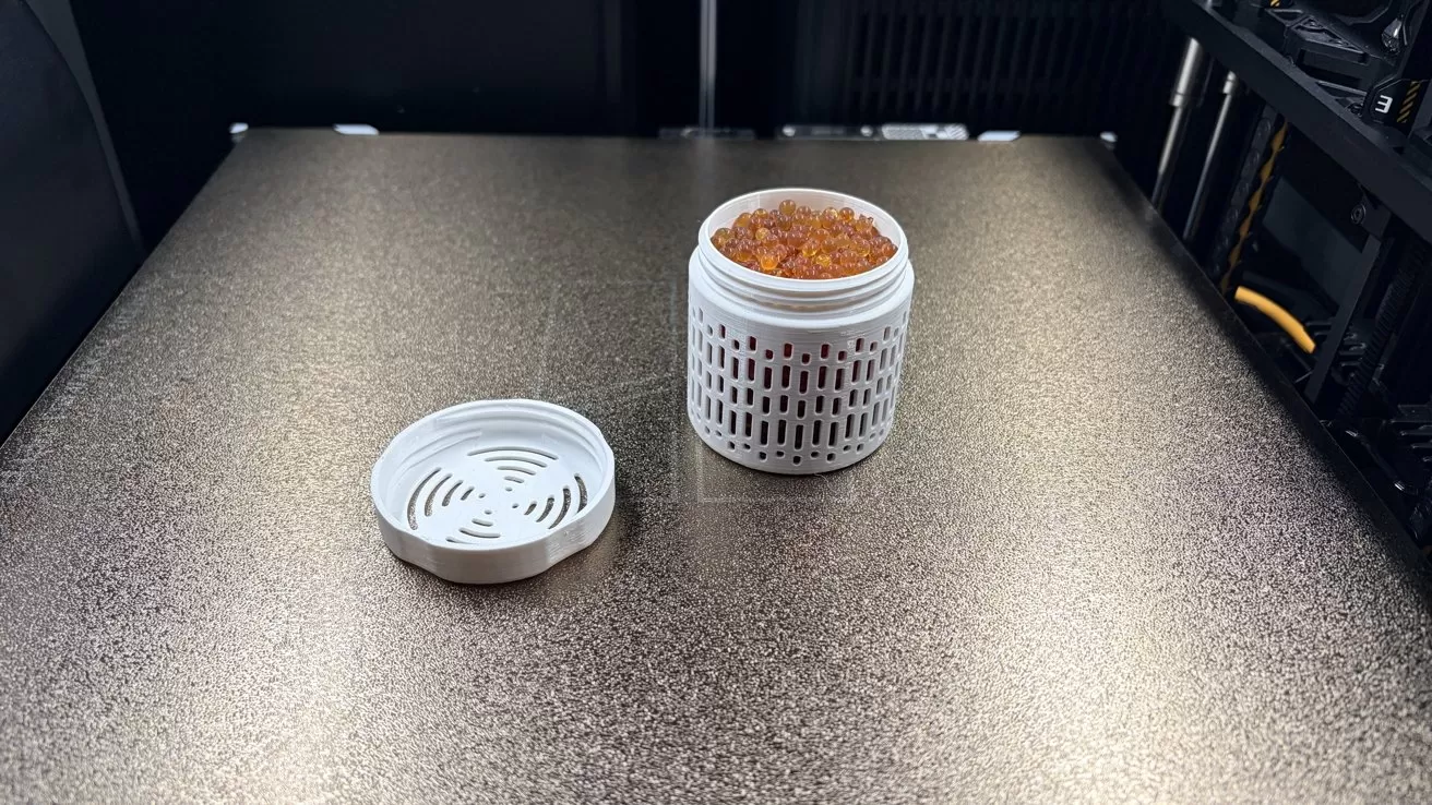 Open white ventilated container filled with small orange beads, with its circular lid lying beside it on a textured metallic surface inside a 3D printer enclosure