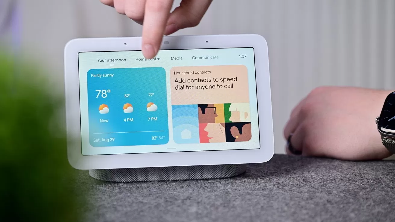 Person interacting with a white smart display on a table, showing a weather forecast of 78 degrees and colorful contact tiles, with a smartwatch visible on the person's wrist