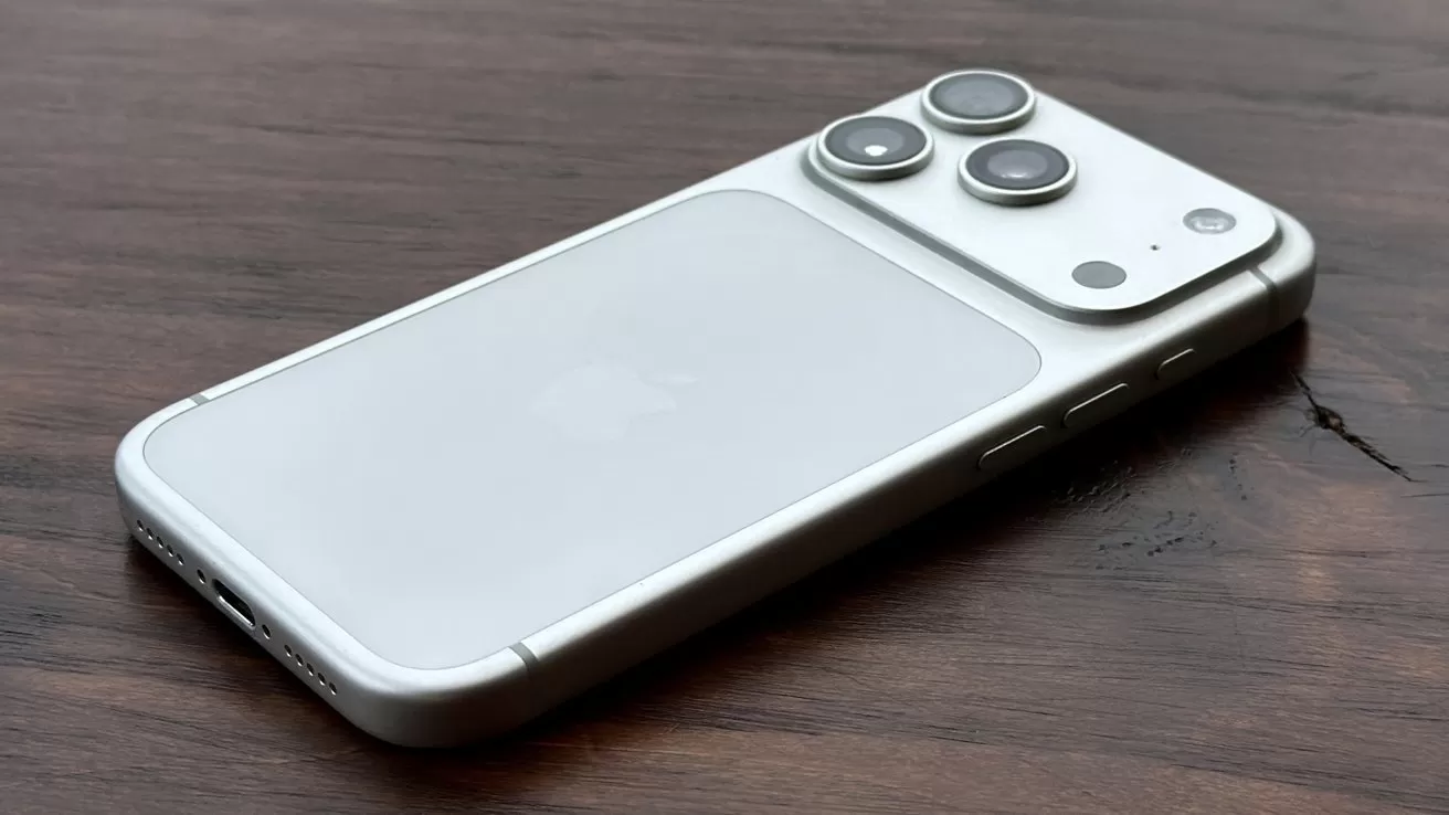 Silver iPhone lying face down on a dark wooden surface, showing a raised triple camera module, side buttons, and the faint Apple logo on the back