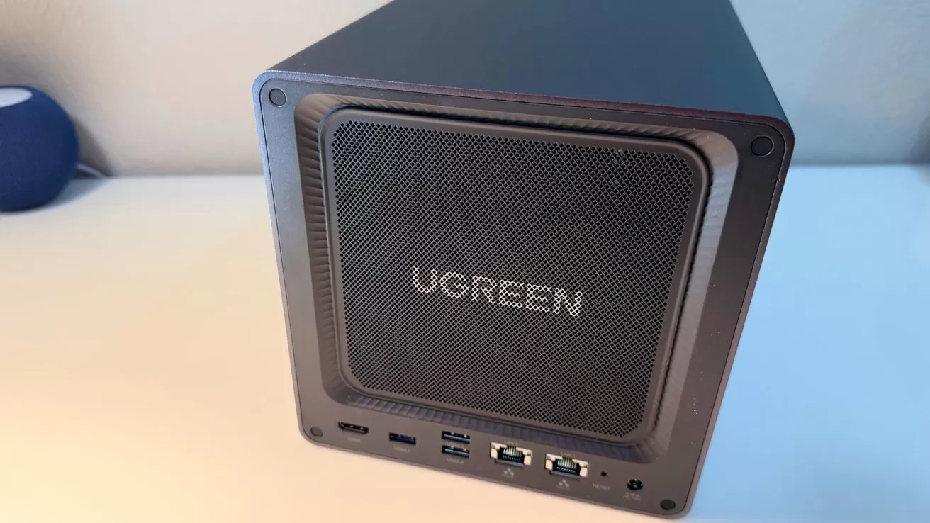 Black UGREEN cube-shaped electronic device on a white desk, front mesh panel with logo, multiple USB and audio ports along the bottom edge, slightly angled perspective