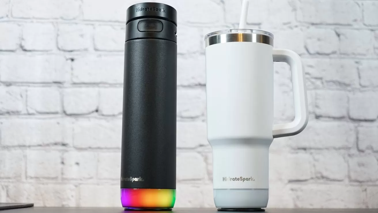 Two HidrateSpark water bottles on a table, one black with a rainbow base and one white with a handle and straw, against a white brick wall background.