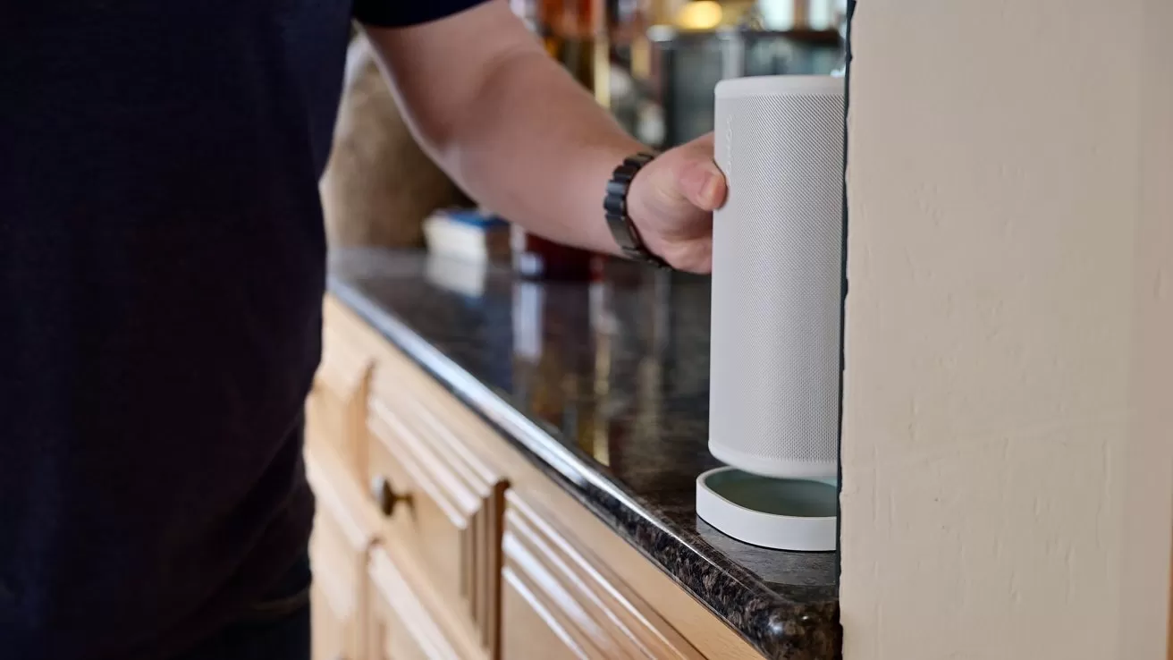 Person placing a tall white smart speaker or router on a dark granite kitchen countertop beside wooden cabinets, with only their arm and torso visible