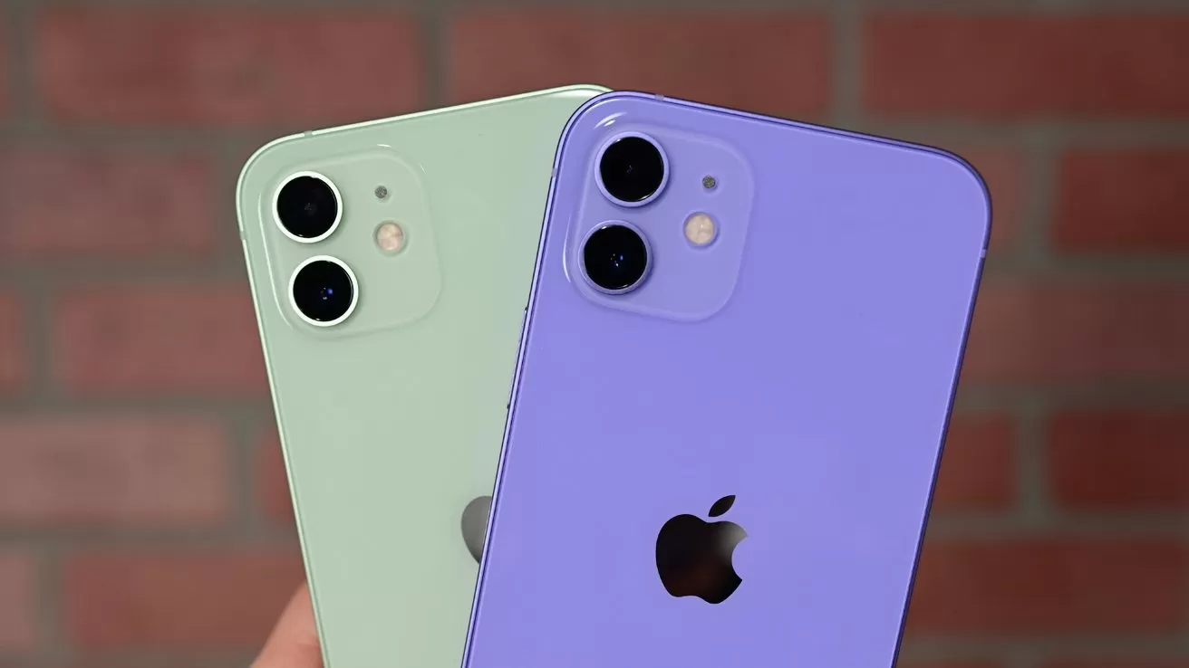 Two iPhones held up, one light green and one purple, both showing their backs with dual cameras and Apple logos, against a blurred red brick wall background