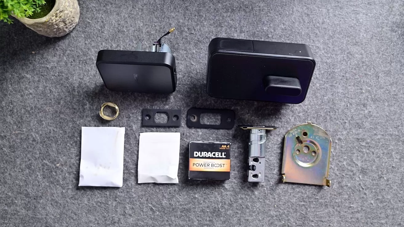Electronic door lock components laid out on gray surface, including black lock units, metal latch and strike plates, screws in packets, mounting hardware, and a Duracell battery, with plant partially visible.
