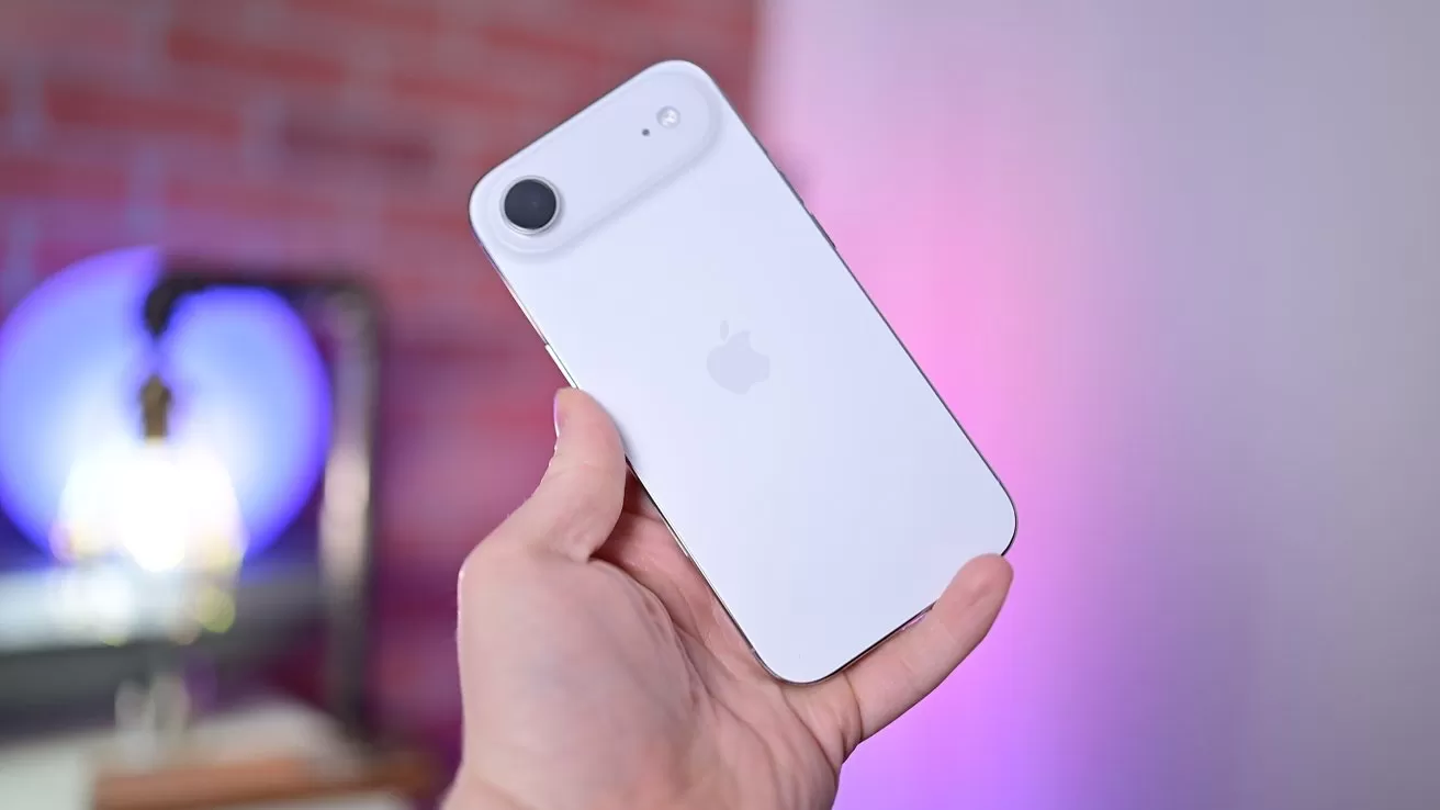Hand holding a white iPhone with a single rear camera against a softly blurred background featuring pink and purple lighting and a partially visible brick wall