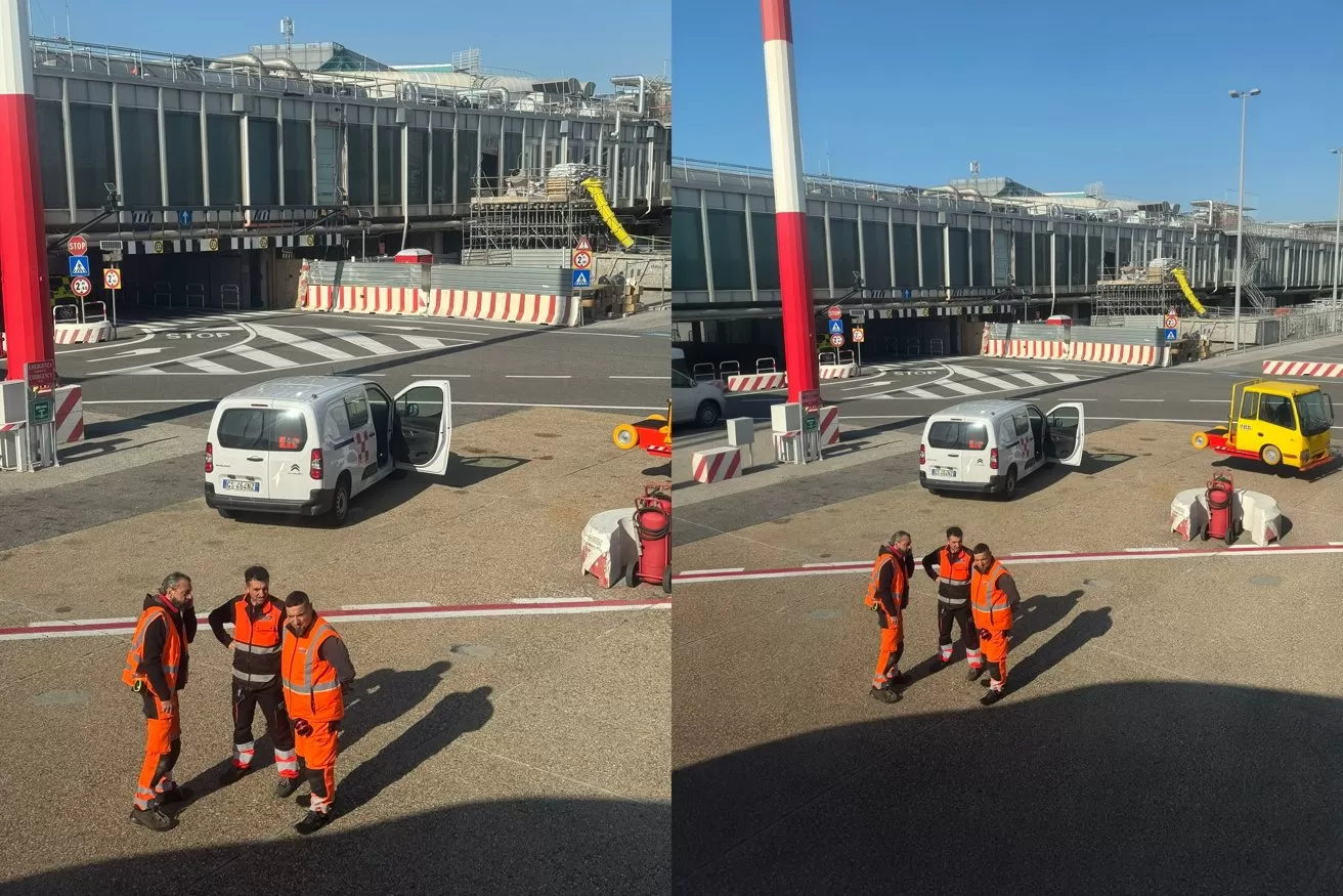 Two airport ground crew in orange uniforms walk on the tarmac near a white van, baggage carts, and terminal buildings with jet bridges under a clear blue sky