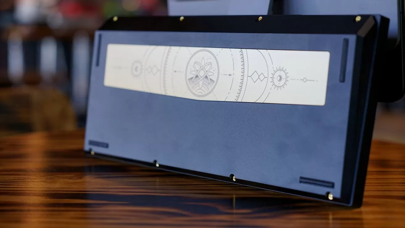 Back of a dark mechanical keyboard with a long cream-colored metal accent plate, engraved with minimalist celestial and geometric designs, resting on a polished wooden desk