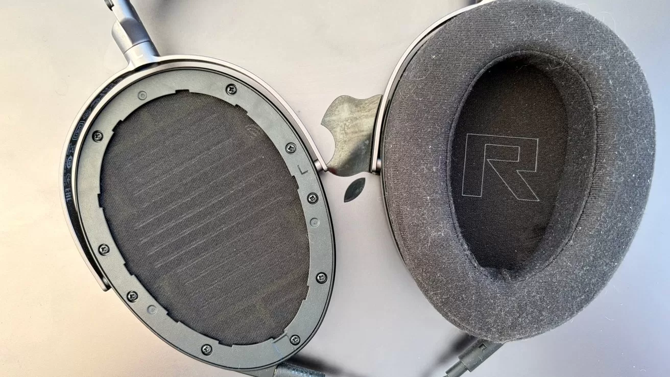 Close-up of over-ear headphones with one ear pad removed, revealing the internal speaker, and the other pad labeled R for right, resting on a light surface
